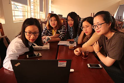 A young woman showing something to four female students. They all sitting around the table. 
